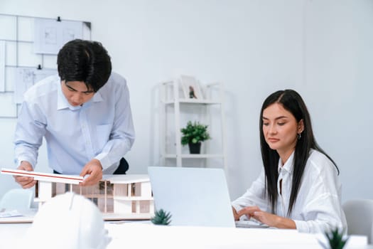 Professional male asian architect using ruler to measure house model length while young beautiful caucasian colleague using laptop to analyzed data on meeting table with house model. Immaculate.