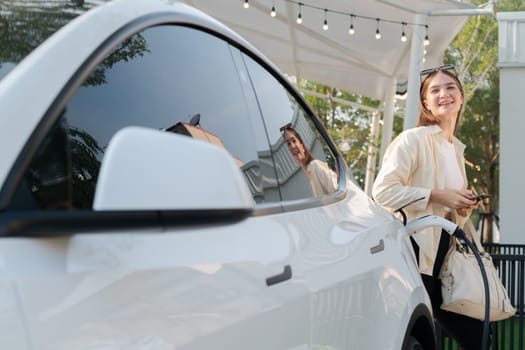 Sustainable urban commute with young woman pay electricity for EV electric car recharging at outdoor cafe in springtime garden, green city sustainability and environmental friendly EV car. Expedient