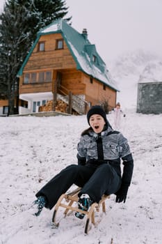 Little girl stands near the house and looks at her screaming mom going down the hill on a sled. High quality photo