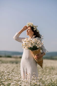 Happy woman in a field of daisies with a wreath of wildflowers on her head. woman in a white dress in a field of white flowers. Charming woman with a bouquet of daisies, tender summer photo.