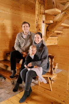 Smiling dad sitting on the stairs next to mom and little girl sitting on a chair in a wooden cottage. High quality photo