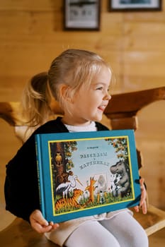 Little smiling girl sits on a chair with a colorful book in her hands. Title: Stories in Pictures. High quality photo