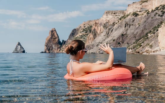 Woman freelancer works on laptop swimming in sea on pink inflatable ring. Pretty lady typing on computer while floating in the sea on inflatable donut at sunset. Freelance, remote work on vacation