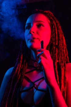 Caucasian girl with dreadlocks smokes a vape in red blue light