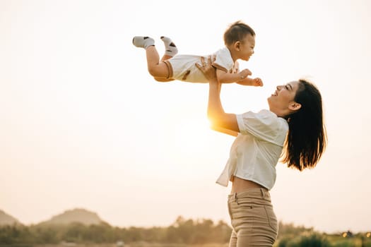 A woman holds her newborn baby up high, as they enjoy a moment of nature in the park at sunset. The little one looks up at the sky with wonder, while the proud mom captures the precious life moment
