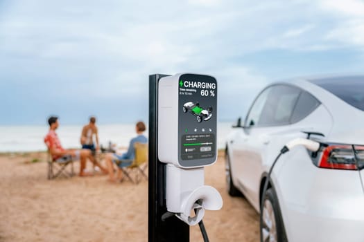 Alternative family vacation trip traveling by the beach with electric car recharging battery from EV charging station with blurred family enjoying the seascape background. Perpetual