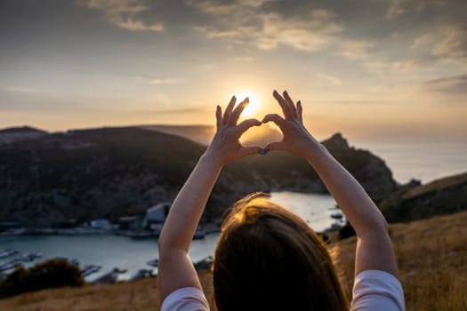 Happy woman on mountain peak, she makes heart shape with hands. Mountain, overlooking sea at sunset. Depicting love shape amidst scenic natural setting