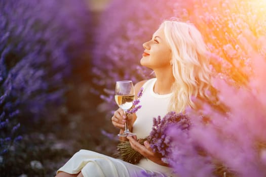 Blonde lavender field holds a glass of white wine in her hands. Happy woman in white dress enjoys lavender field picnic holding a large bouquet of lavender in her hands . Illustrating woman's picnic in a lavender field