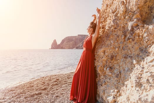 Side view a Young beautiful sensual woman in a red long dress posing on a rock high above the sea during sunrise. Girl on the nature on blue sky background. Fashion photo.