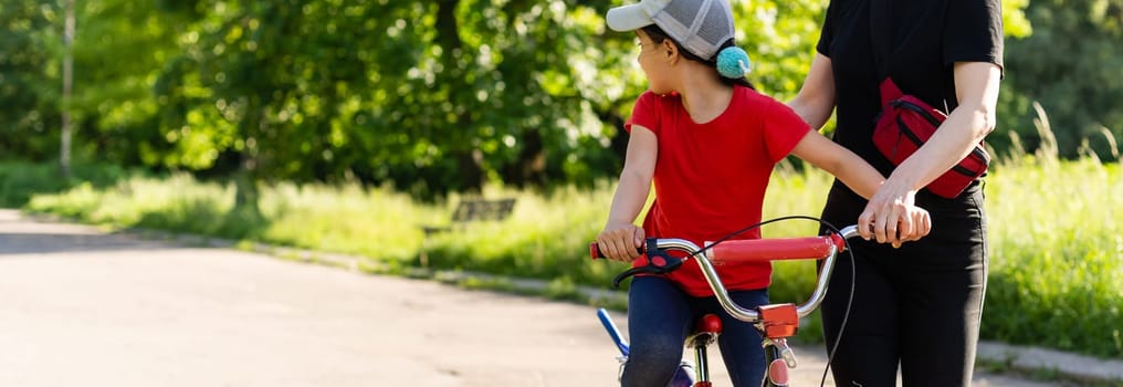 Beautiful and happy young mother teaching her daughter to ride a bicycle. Both smiling, summer park in background