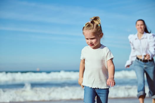 Pensive blonde little child girl 2-3 years old in white t-shirt and casual denim, walking on the beach against blurred background of her happy mother running barefoot, enjoying wonderful time together