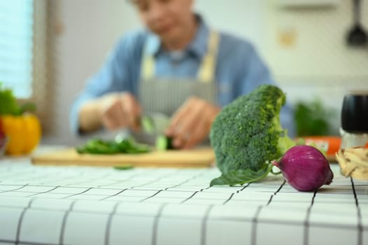 Select focus fresh vegetables on kitchen table with blurred senior man cooking in background.