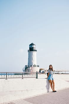 Young woman stands leaning on a concrete fence near the lighthouse and looks to the side. High quality photo