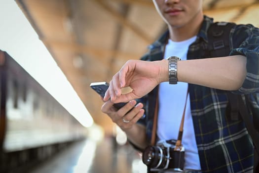 Male traveler holding mobile phone and checking time on wrist watch while waiting for her train at platform.