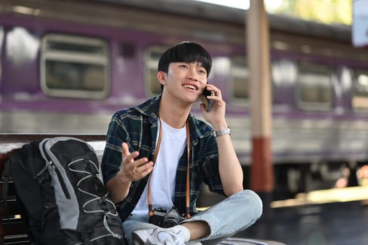 Cheerful young male traveler talking on mobile phone while waiting for the train at the train station.