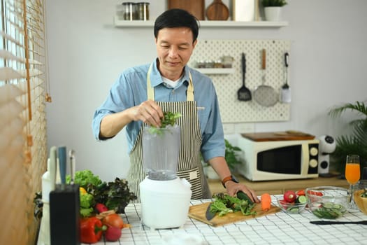 Happy middle age man making vegetable smoothies with blender in kitchen. Healthy lifestyle concept.
