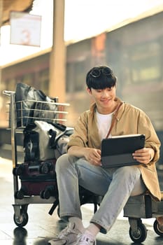 Smiling young man sitting on luggage cart and using digital tablet, waiting for the train.