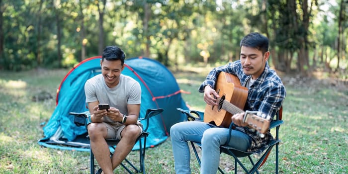 LGBTQIA Gay couple camping together in woods for holidays and relax on guitar together.