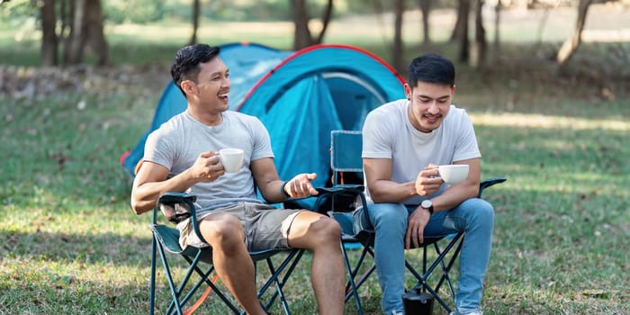 Gay LGBTQIA couple sitting on picnic chair drinking tea and coffee while camping on vacation holiday.