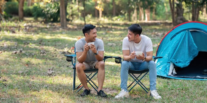 Gay LGBTQIA couple sitting on picnic chair drinking tea and coffee while camping on vacation holiday.