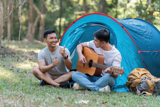 LGBTQIA Gay couple camping together in woods for holidays and relax on guitar together.