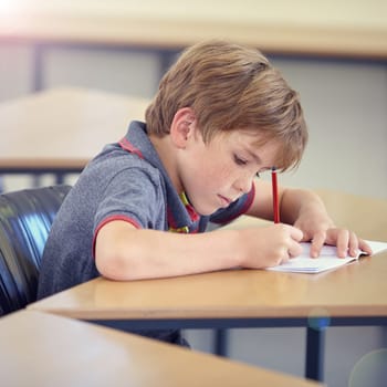 Child, student and writing in classroom for learning, education and development in language quiz or test. Smart boy or kid with notebook for school progress, creativity and knowledge at his desk.