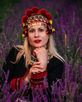 A girl in a chelsea headdress and a black dress decorated with red embroidery sits between lavender bushes, Ukrainian traditional and national clothing.