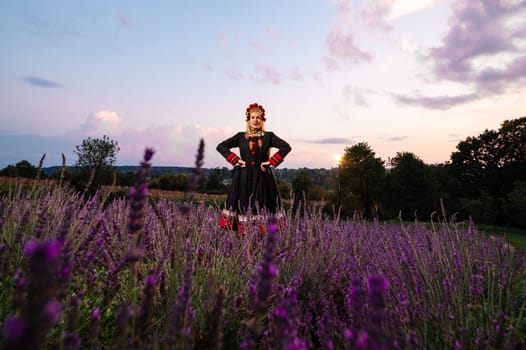 A girl in a chelsea headdress and a black dress decorated with red embroidery stands between lavender bushes.