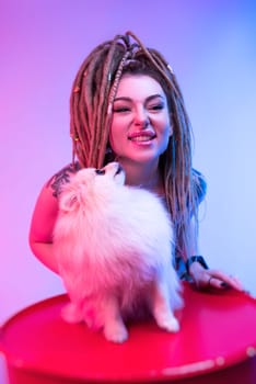 cool girl with braided dreadlocks on her head with a cute Pomeranian dog in neon light on a light background