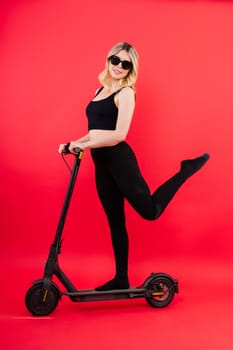 Female in a sport clothes stands on electric scooter with sunglasses on red and white background