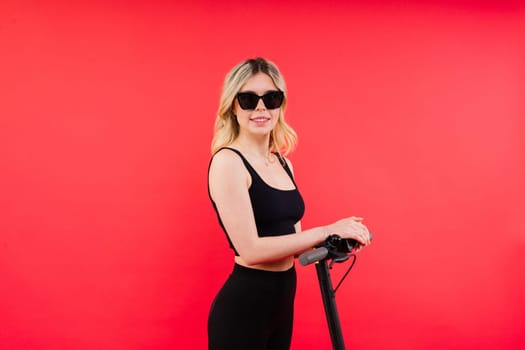 Female in a sport clothes stands on electric scooter with sunglasses on red and white background