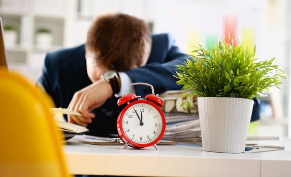 Red alarm clock shows late time closeup and tired office male clerk in suit take nap on table workplace full of exam papers. Career frustration freelance employment fail study problem low energy down