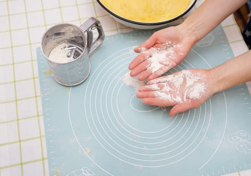 The image shows hands covered in flour over a silicone baking mat. A sifter and mixing bowl are nearby