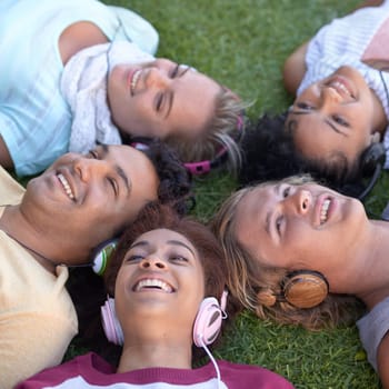 Friends, headphones and group relax on grass at park, listening to music or radio in summer. Audio, smile and students on lawn in top view, nature and young people streaming sound together outdoor.