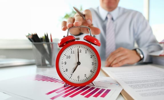 Male hand on the alarm clock a red color stands in the office on the table showing seven o'clock in the morning or evening AM PM.