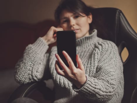 A caucasian young beautiful girl with brown hair and in a thick gray knitted sweater sits in her bedroom on an office chair and chats online with her friend holding a black smartphone in front of her and smiling supports her face with her hand, side view close-up with depth of field. Online chatting concept , social media, modern lifestyle.