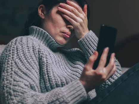A caucasian young beautiful girl in a thick gray knitted sweater sits in her bedroom on the floor cuddled up to the bed and chats online with her friend, covering her face with her palm from bad news, holding a black smartphone in front of her, bottom view close-up with depth of field. Online chatting concept, social media, modern lifestyle, psychology.