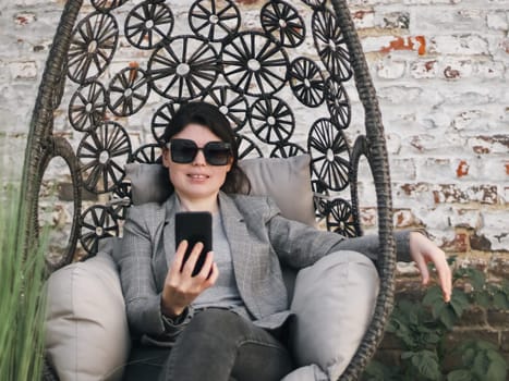A young beautiful caucasian girl in a plaid jacket, gray t-shirt, jeans, sunglasses holds a phone in her hands and looks at the smartphone screen while smiling, sitting in a hanging chair against the background of an old brick wall in the backyard of her house, close-up side view. Concept using technology, social networks, online chats, modern lifestyle, at home, social media.