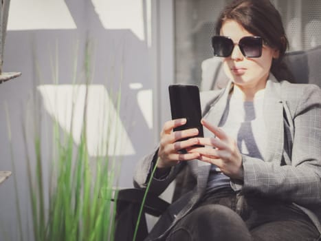 Young beautiful caucasian girl in a plaid jacket, gray t-shirt, jeans and sunglasses holds a smartphone in front of her while sitting in a garden chair in front of a window in the backyard of her house, close-up side view. Concept using technology, social networks, social media, online chats, modern lifestyle, home, online business conference.
