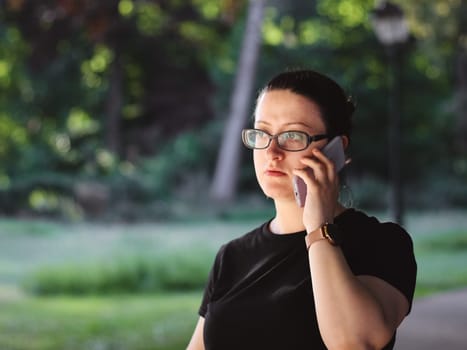 Caucasian woman in glasses and a black t-shirt emotionally talking on a mobile phone waving her hand in a public park against the background of blurry trees, close-up side view. Concept of using a mobile phone, online chatting, social media, using technology.