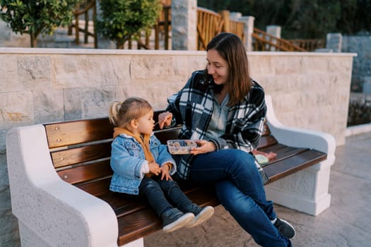 Smiling mom feeds a little girl from a spoon while sitting on a bench. High quality photo