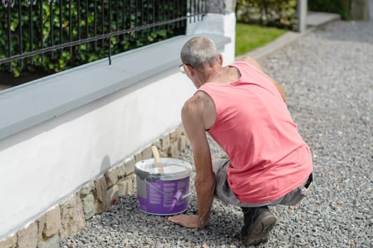 A middle-aged man paints a fence with white paint with a brush, repairs the damaged surface, High quality photo