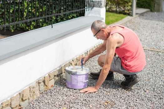 A middle-aged man paints a fence with white paint with a brush, repairs the damaged surface, High quality photo