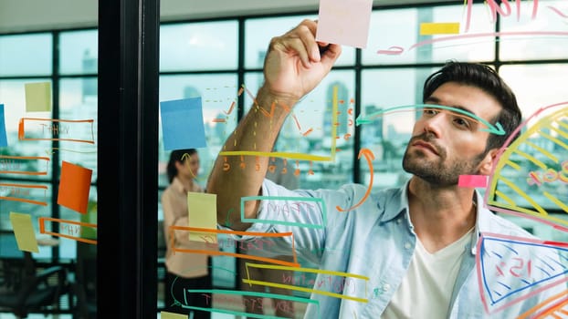 Professional businessman uses sticky notes at glass wall with graph to brainstorming idea while multicultural businesspeople discussing about marketing ideas at office with city view. Tracery