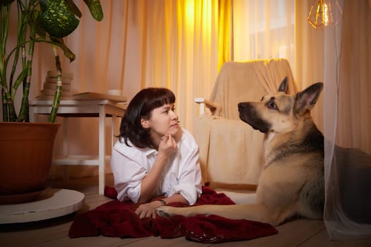 Adult mature woman with big shepherd dog in white shirt. Room with girl and calm cozy evening atmosphere with transparent curtains and soft warm light of lamps. Concept of love for animals, pets