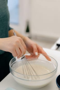 The hand of one young Caucasian unrecognizable girl mixes the dough manually with a whisk in a glass bowl, standing at a white table, close-up view from below. Step-by-step instructions for baking synabons. Step 2.