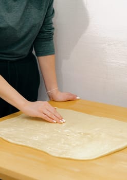One unrecognizable young Caucasian girl spreads butter with her fingers onto the yeast dough on a wooden table, standing in the kitchen, close-up side view. Step-by-step instructions for baking synabons. Step 6.