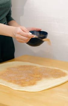 One unrecognizable young Caucasian girl sprinkles brown sugar with cinnamon onto the yeast dough on a wooden table using a teaspoon, standing in the kitchen, close-up side view. Step-by-step instructions for baking synabons. Step 7.