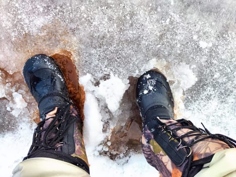 Feet of Hunter or fisherman in big warm boots on winter day on snow. Top view. Fisherman on the ice of a river, lake, reservoir on spring day with melting ice. Dangerous fishing in autumn