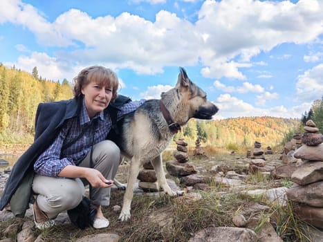 Girl with shepherd dog taking selfie near water of mountain river or lake with stones. Middle aged woman and big pet on nature. Friendship, love, fun and hugs